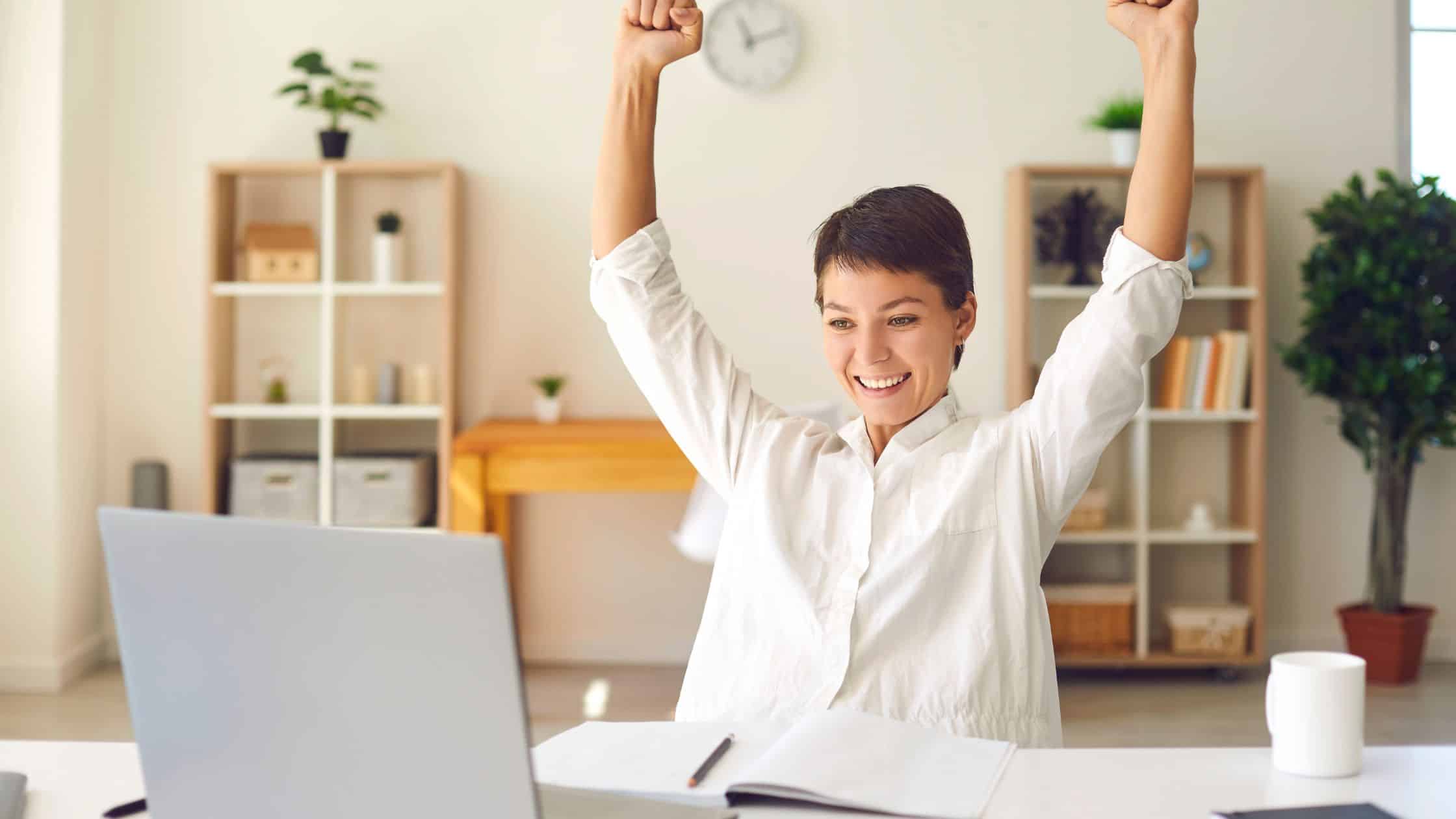 A young woman sat at her desk, triumphantly raising her arms in celebration of her success.